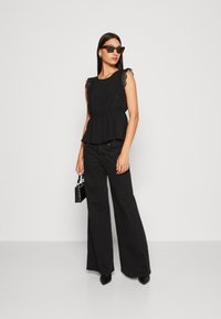 Must have peplum top mustriga pitsi ääristustega õlgadel, kombineeritud mustade laiemate pükstega. Mudel hoiab käes väikest musta käekotti ja kannab tumedaid prille.
