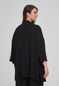 Cardigan oversize nero con maniche ampie e colletto a scialle. Tessuto leggero con una consistenza liscia. Presenta spacchi laterali per una maggiore libertà di movimento.