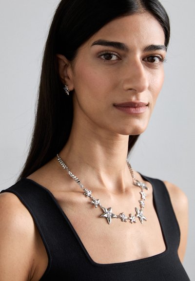 Collier en forme d'étoile argentée avec des accents de strass, porté sur un haut noir. Présente un design de chaîne liée avec des tailles d'étoiles variées.