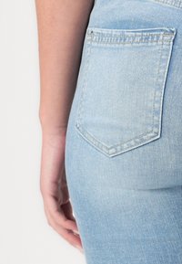 Világoskék farmernadrág, látható hátzsebbel, amely hegyes, szögletes formájú. Az anyag sima, enyhén strukturált megjelenésű.