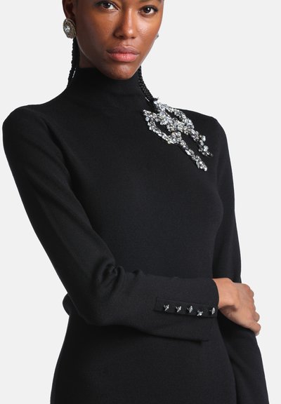Femme portant un haut noir à manches longues avec col montant, décoré d'embellissements en strass sur l'épaule et le poignet de la manche.