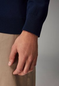 Tengerészeti kötött pulóver bordás mandzsettával, világos színű anyag keretezi. Közeli felvétel egy kézről, amely a szöveten pihen, bemutatva a textúrát.