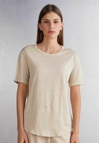 Béžové tričko s krátkým rukávem vyrobené z lehkého, texturovaného materiálu s kulatým výstřihem a mírně zakřiveným lemem.
