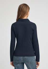 Marinefarbener Langarm-Polo mit Kragen, taillierte Passform, glatte Textur, kombiniert mit hellblauen, destroierten Jeans und einem dezenten Rücken-Detail.