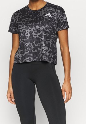 Osoba nosící černo-šedé tričko Adidas s maskáčovým vzorem v kombinaci s černými přiléhavými legínami, stojící před jednoduchým pozadím.