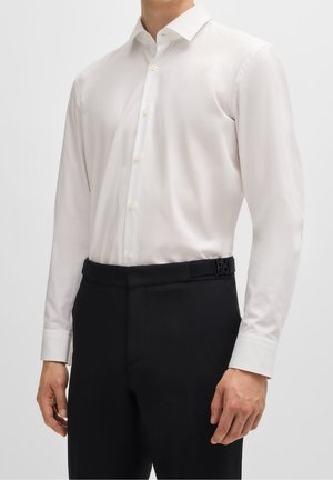 Mężczyzna ubrany w dopasowaną, białą koszulę na guziki, włożoną do czarnych, dopasowanych spodni, na jednolitym tle.