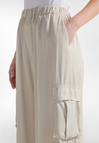 Beige elastische taillebroek gemaakt van lichtgewicht stof. Voorzien van zijzakken en een cargozak met klep. Soepele textuur, losse pasvorm.