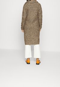 Dlouhý pletený kabát v hnědých tónech s texturovaným vzorem. Kombinován s bílými rozšířenými kalhotami a oranžovými botami. Minimalistický design, s kapucí.