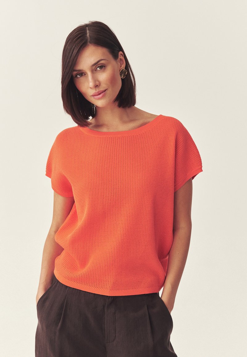 Femme aux cheveux courts et foncés portant des boucles d'oreilles en or, un haut orange texturé à manches courtes et un pantalon foncé, debout avec les mains dans les poches.