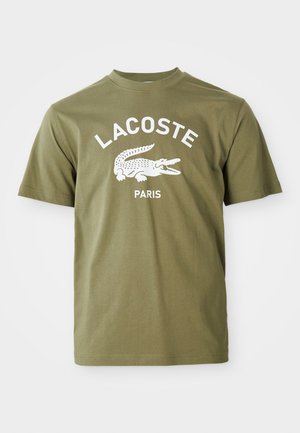 Camiseta de algodón verde oliva con un gráfico de cocodrilo blanco y el texto "LACOSTE PARIS", con cuello redondo y mangas cortas.