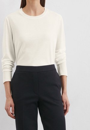 Frau trägt einen eng anliegenden weißen Langarmpullover, der in eine hoch taillierte schwarze maßgeschneiderte Hose gesteckt ist, vor einer schlichten hellen Wand.