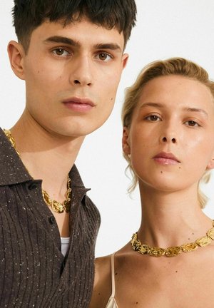 Man en vrouw dragen bijpassende chunky gouden kettingen. De vrouw heeft een beige top met dunne bandjes aan, de man een donkere textuurshirt met een wit onderhemd.