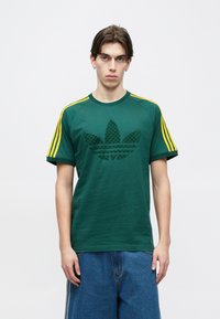 Jauns vīrietis zaļā Adidas krekliņā ar dzeltenām svītrām uz piedurknēm un džinsiem, stāvot pret balto vienkāršo fonu.