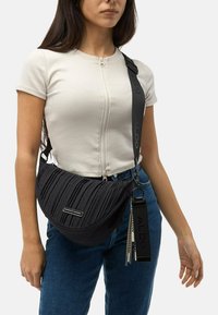 Černá ramenní taška s plisé texturou, stříbrnými detaily a nastavitelným popruhem. Nositá přes rameno k casual outfitu.