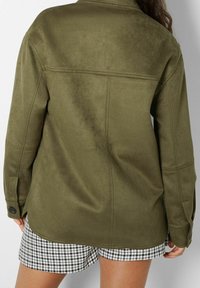 Giacca camicia verde oliva con una finitura morbida e testurizzata, con maniche lunghe e polsini con bottoni. Indossata sopra pantaloni corti a scacchi.