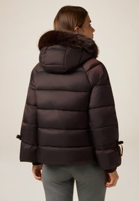 Chaqueta acolchada de color marrón oscuro con capucha de piel sintética, que presenta secciones acolchadas horizontales y puños ajustables con lazo. Textura suave.