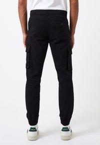 Pantalones cargo negros con puños elásticos, bolsillos laterales y una tela texturizada. Llevados con zapatillas blancas con detalles en verde.