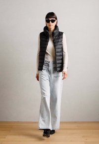 Gilet matelassé noir, col roulé ajusté et jean délavé à jambes larges. Le mannequin porte des lunettes de soleil sombres et des chaussures noires, sur un sol en bois.
