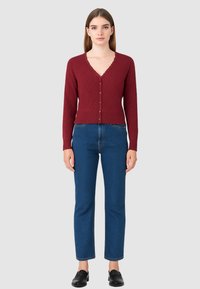 Donna che indossa un cardigan a maglia bordeaux con bottoni, jeans blu a gamba dritta e scarpe nere senza lacci, in piedi su uno sfondo grigio neutro.