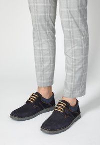 Navy blue casual shoes with a textured finish, featuring brown laces, contrasting stitching, and gray rubber soles, paired with plaid trousers.