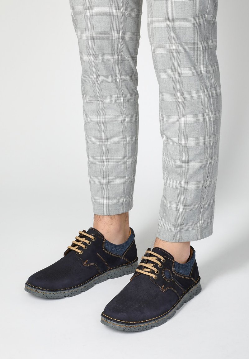 Navy blue casual shoes with a textured finish, featuring brown laces, contrasting stitching, and gray rubber soles, paired with plaid trousers.