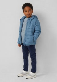 Hellblaues Puffer-Jacket mit Kapuze, grauer Strickpullover, marineblaue Hose und weiße Sneakers. Die Jacke verfügt über einen Reißverschluss und ein gestepptes Design.