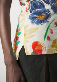 Top de tela ligera con estampado floral de flores coloridas, aberturas laterales en el dobladillo, presenta acentos en azul marino, amarillo, naranja y verde.