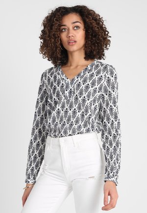 Camisa de manga larga en blanco y azul marino con patrón floral, escote en V y puños fruncidos, combinada con vaqueros blancos desgastados.
