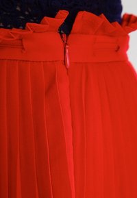 Primer plano de una falda roja plisada con una cintura con volantes y un cierre de cremallera centrado.
