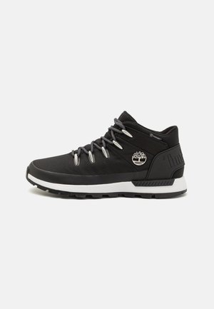 Zapatilla de media caña negra con parte superior de tela texturizada, ojales de metal, cordones grises y media suela blanca; presenta un logotipo de árbol en el lateral.
