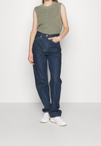 Top de punto sin mangas verde combinado con jeans de mezclilla oscuros de tiro alto y zapatillas blancas. Ajuste holgado con detalles de bolsillos visibles.