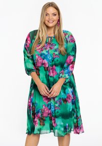 Bloemenjurk van groene stof met roze en blauwe accenten. Heeft een losse pasvorm, lange mouwen en een strikdetail bij de hals. Lichtgewicht en vloeiende ontwerpen.