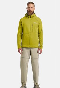 Groen fleecejack met rits en capuchon, voorzien van twee voorzakken, gecombineerd met lichtbeige afritsbare broek. Geschikt voor buitenactiviteiten.