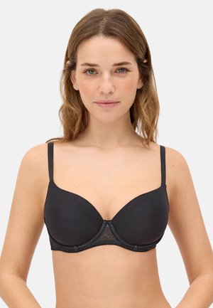 Mujer con cabello castaño hasta los hombros, llevando un sujetador negro con aro y tirantes ajustables finos, mirando hacia adelante sobre un fondo blanco.