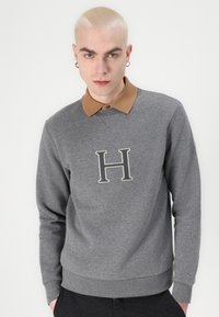 Grauer Hoodie mit braunem Kragen, der auf der Brust mit einem gestickten Buchstaben "H" verziert ist. Glatte Textur, taillierte Passform und gerippter Bund und Bündchen.