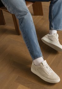 Beige Wildlederschuhe mit weißen Gummisohlen, kombiniert mit hellblauen Jeans und weißen Socken, auf einem Holzboden.