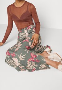 Haut marron à manches longues, avec des panneaux transparents au niveau de la poitrine. Jupe verte fluide avec des motifs floraux roses et des papillons. Chaussures blanches à talons carrés.