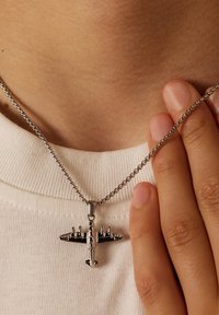 Zilveren metalen halsketting met een vintage vliegtuig hanger, met gedetailleerde accenten en een ronde schakelketting, rustend op een lichte stoffen achtergrond.