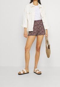 White denim jacket, white T-shirt, floral purple shorts, and brown sandals with dual straps, holding a woven bag.