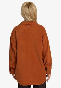 Rust-colored fleece shirt with a collared neck, oversized fit, and buttoned cuffs. Soft texture and a subtle brand tag on the back.