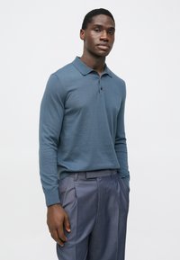 Polo azul de manga comprida feita de tecido liso, com gola clássica e pala com dois botões, combinada com calças cinzentas pregueadas.