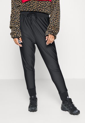 Persona vistiendo pantalones jogger negros ajustados con cintura de cordón, camiseta de manga larga con estampado de leopardo y zapatos deportivos negros.