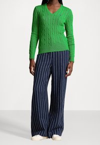 Suéter verde de punto trenzado con cuello en V, mangas largas, combinado con pantalones de pierna ancha a rayas en azul marino y blanco, y zapatillas blancas.