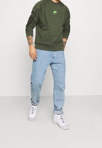 Green sweatshirt with a small white Nike Air logo, paired with light blue denim jeans and white high-top sneakers. Casual fit and style.