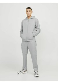 Sudadera gris con cremallera, capucha y bolsillos frontales, combinada con pantalones de chándal ajustados a juego. Ambos fabricados en un tejido suave y con un diseño minimalista.