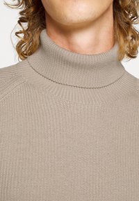 Nahaufnahme einer Person, die einen beigen gerippten Rollkragenpullover trägt, wobei der Hals und lockige Haarsträhnen zu sehen sind.