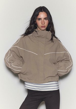 Femme aux longs cheveux noirs portant une veste ample couleur beige avec des liserés blancs, un chemisier rayé en dessous, et un pantalon noir, debout devant un fond uni.