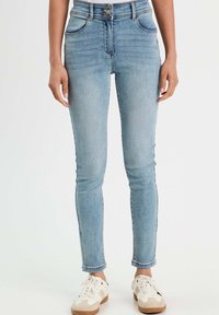Jean taille haute bleu clair avec une coupe slim, comportant cinq poches, des passants de ceinture et une fermeture à bouton. Porté avec des baskets blanches.