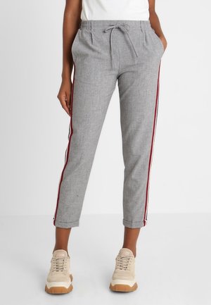 Persona que lleva pantalones grises con cordón y franjas laterales rojas y blancas, y zapatillas deportivas beige, de pie frente a un fondo blanco liso.