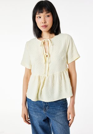 Femme portant un chemisier crème à manches courtes avec une texture florale et des détails à nouer au niveau du col, assorti à un jean en denim bleu.